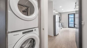 A laundry room with a front-loading washer and dryer, a kitchen with stainless steel appliances, and a large window providing natural light. - Landing | Spectacular 1BD, Courtyard, Pool (Richmond)