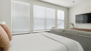 Habitación de hotel con una cama, un sofá gris, un televisor de pantalla plana y ventanas amplias con persianas.