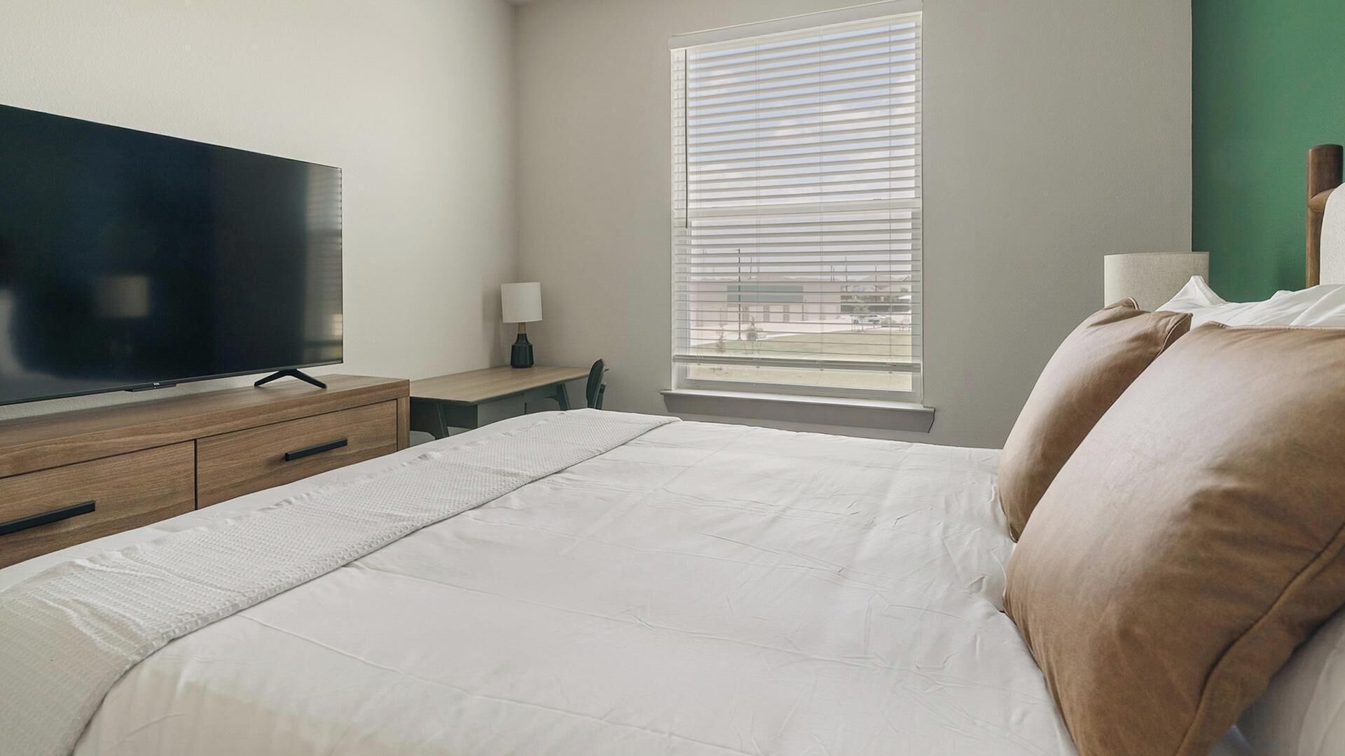 Quarto com televisão de tela plana grande, mesa de cabeceira de madeira, mesinha de cabeceira com abajur, janela com persianas e cama com roupa de cama branca e duas almofadas.
