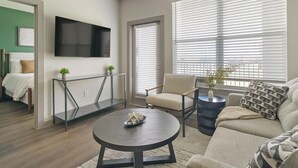 En moderne stue med sofa, lænestol, sofabord, fjernsyn og et stort vindue med persienner.