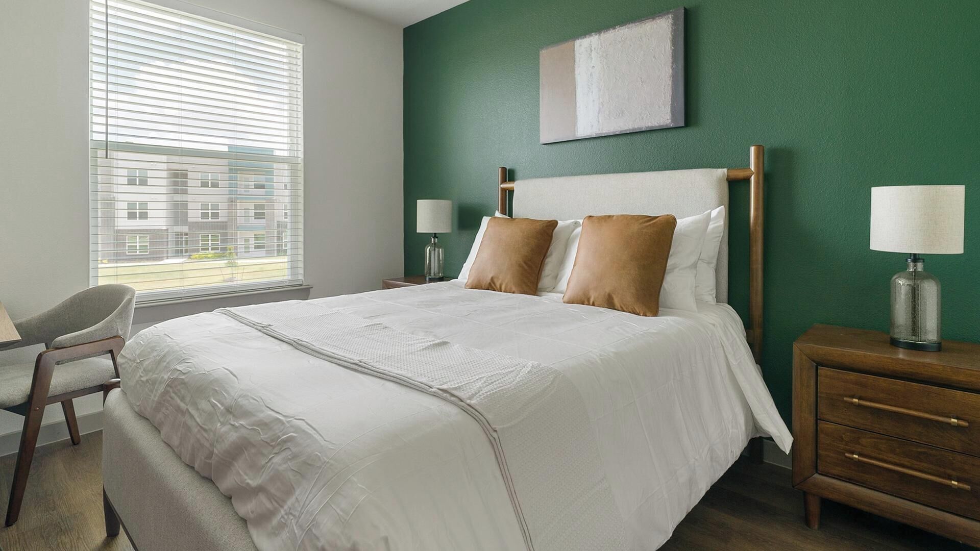A hotel room with a bed, a chair, a nightstand, and a window with blinds.