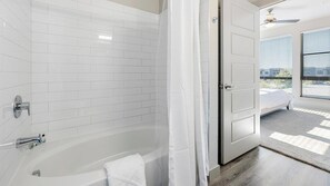 Ein modernes Badezimmer mit Badewanne, weißen Fliesenwänden und einer Tür zu einem Schlafzimmer mit Aussicht.