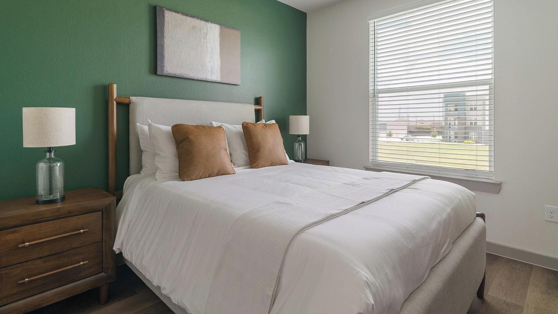 A neatly made bed with white linens and two brown pillows, a bedside table with a lamp, and a window with blinds.