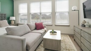 Ein modernes Schlafzimmer mit einem grauen Sofa, einem kleinen weißen Beistelltisch, einer grauen Kommode und einem großen Fenster mit Jalousien.
