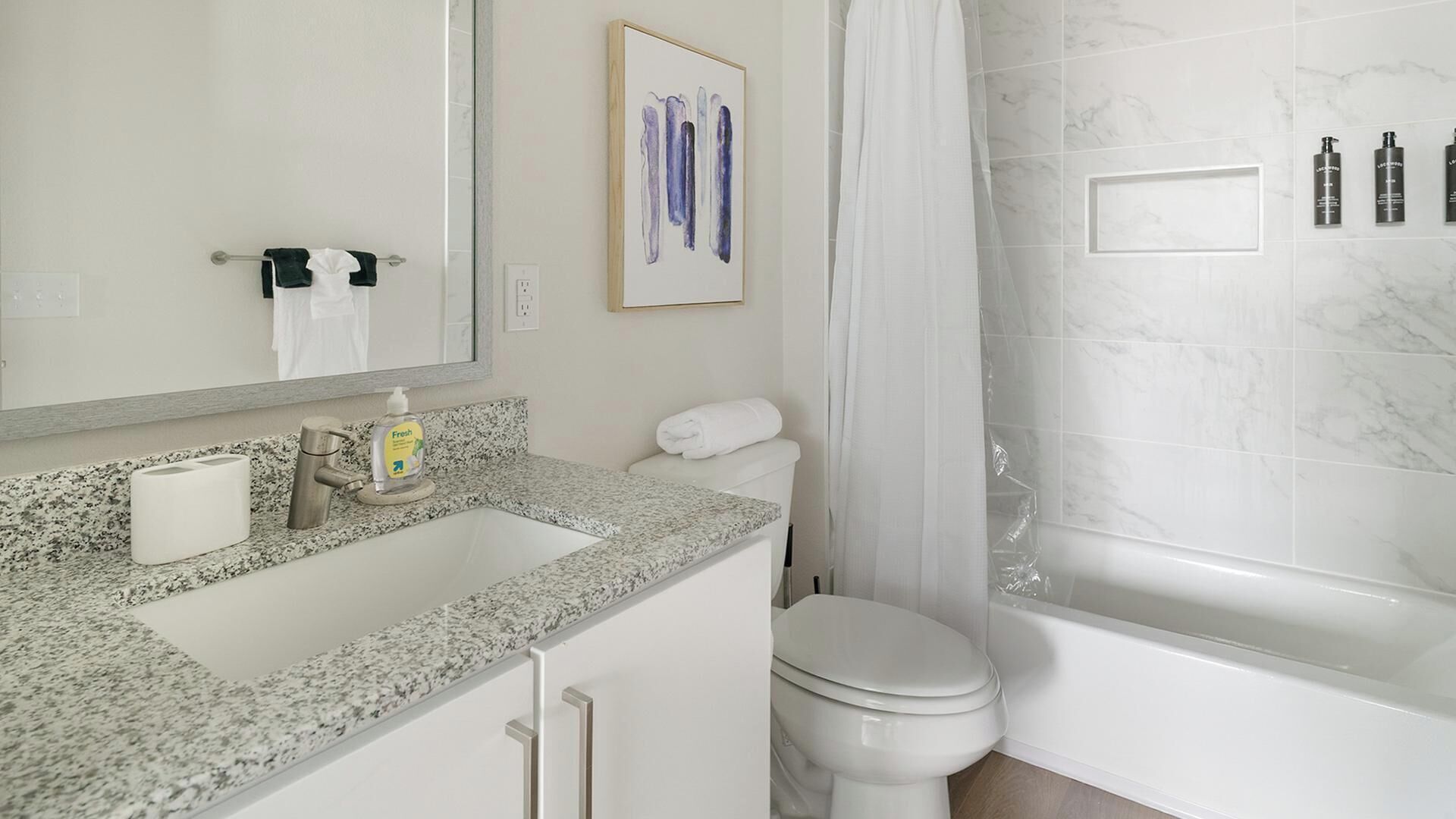 Une salle de bain avec un lavabo sur un plan de travail en granit, des toilettes, une baignoire et un tableau abstrait encadré accroché au mur.