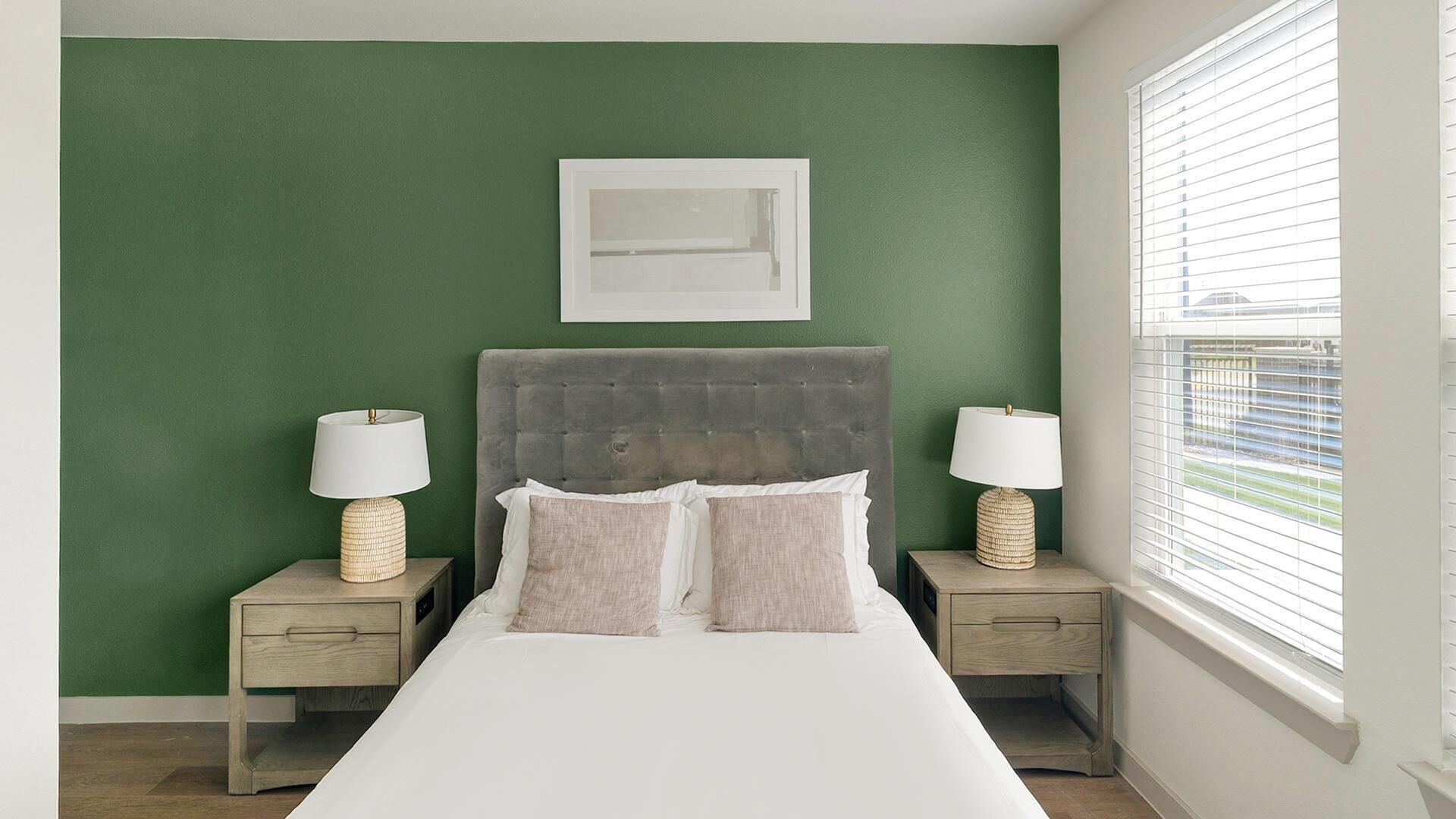 Une chambre avec un mur vert, une tête de lit grise à motifs, deux tables de chevet avec des lampes et une fenêtre avec des stores.