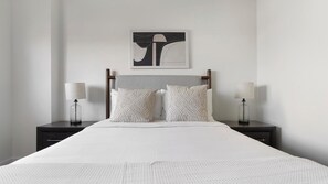 Ein Bett mit weißer Bettdecke, zwei Nachttische mit Lampen und ein an der Wand hängendes abstraktes Kunstwerk.