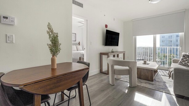 A modern living room with a dining table, a sofa, a TV mounted on the wall, and a large window offering a city view. - Landing | Stylish 1BD, Gym (Hialeah)