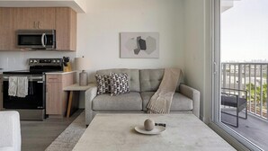 Un espace de vie moderne comprenant une cuisine, un canapé gris agrémenté de coussins à motifs, une table basse ornée d’un objet décoratif et une grande fenêtre donnant sur un balcon.