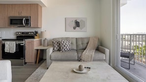 A modern living space with a kitchen, a grey sofa with patterned cushions, a coffee table with a decorative item, and a large window with a balcony view. - Landing | Stylish 2BD, Gym (Hialeah)
