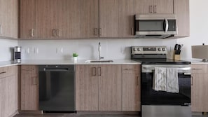 A modern kitchen with wooden cabinets, a stainless steel dishwasher, a microwave, and an oven. - Landing | Stylish 2BD, Gym (Hialeah)