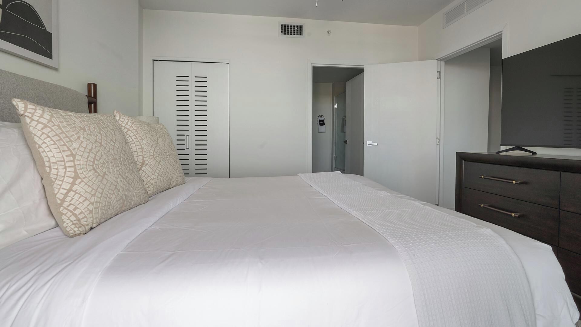 Een slaapkamer met een bed, een dressoir, een spiegel en een deur die naar een andere kamer leidt.