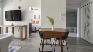 Ett modernt hotellrum med ett matbord i trä, en tv som är monterad på väggen, en säng med vita och orange sängkläder, en liten växt på bordet och ett badrum som syns genom en öppen dörr.