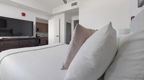A modern bedroom with a large bed, a flat-screen TV, a kitchen area, and a door leading to another room. - Landing | Spectacular 1BD, Gym (Hialeah)
