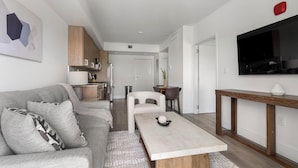A modern living room with a sofa, a coffee table, a TV, and a kitchen area in the background. - Landing | Spectacular 1BD, Gym (Hialeah)