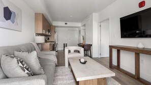 A modern living room with a sofa, a coffee table, a TV, and a kitchen area in the background. - Landing | Spectacular 1BD, Gym (Hialeah)