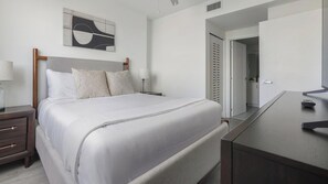 A neatly arranged bedroom with a bed, bedside tables, a lamp, a mirror, and a door leading to a bathroom. - Landing | Stunning 1BD, Gym (Hialeah)