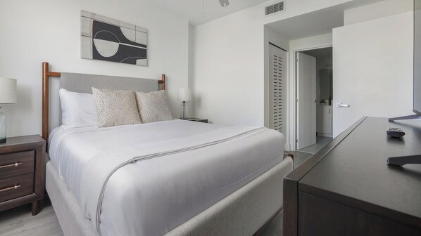 A neatly arranged bedroom with a bed, bedside tables, a lamp, a mirror, and a door leading to a bathroom. - Landing | Modern 1BD, Gym (Hialeah)
