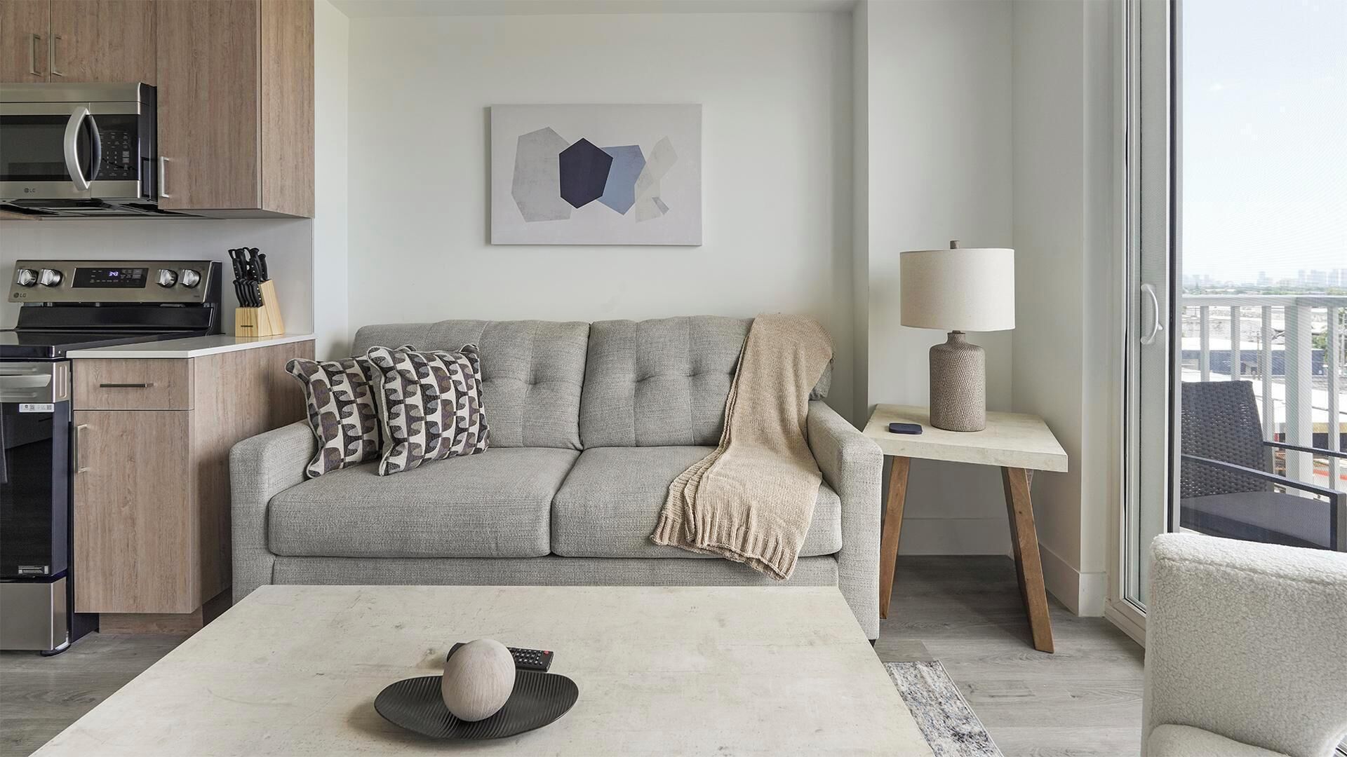 A modern living room with a grey sofa, a coffee table, a lamp, and a kitchen area with a microwave and stove.