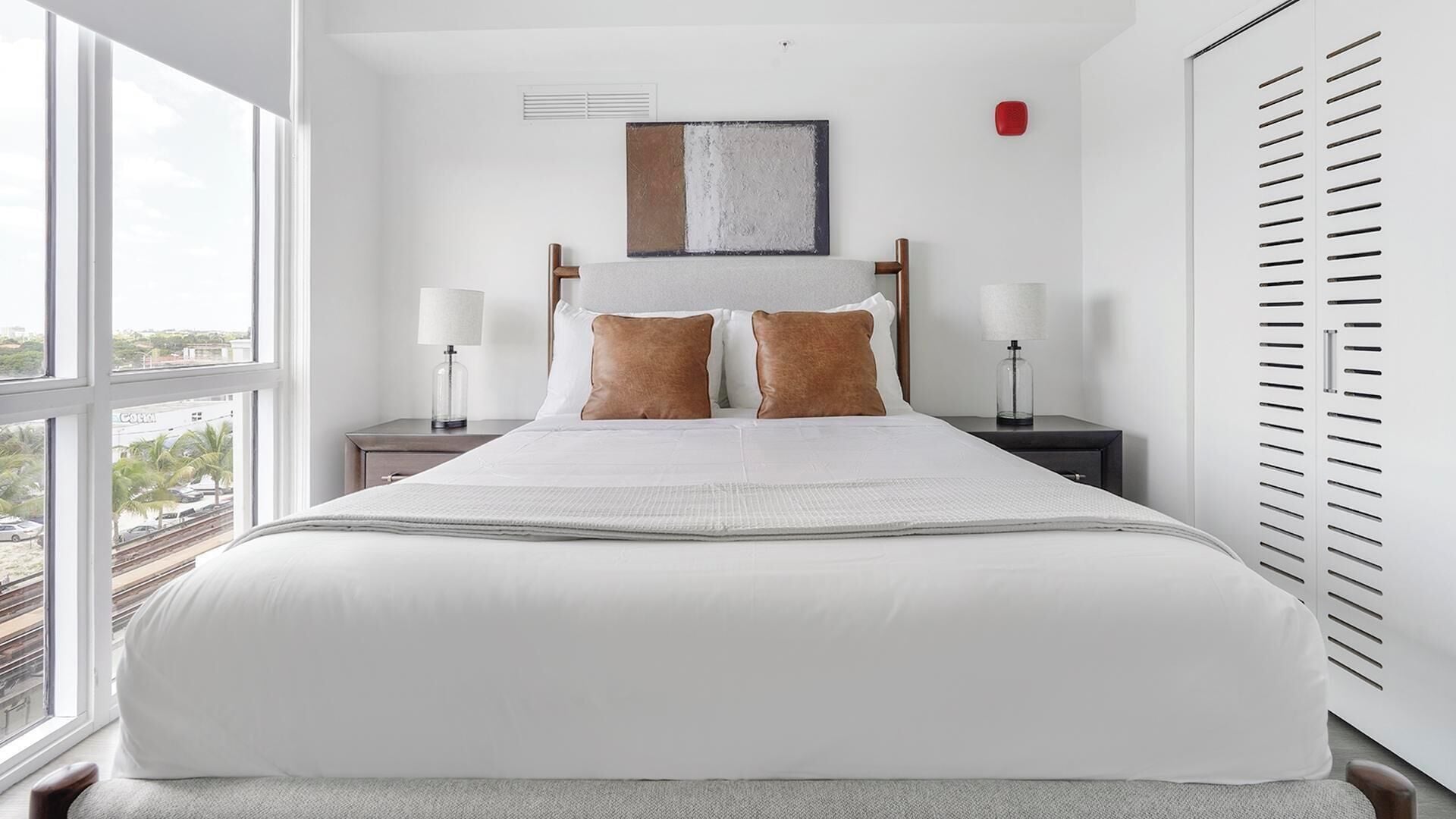 Een hotelkamer met een groot bed, twee nachtkastjes met lampen, een raam met uitzicht, en een rood object aan de muur.