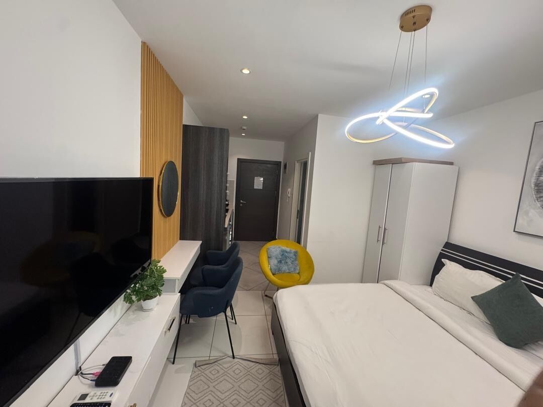 A modern hotel room with a large flat-screen TV, a desk with a plant, a yellow chair, a bed with a green pillow, and a neon light on the ceiling.