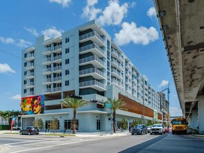 A multi-story hotel with balconies, located at a street corner with palm trees and a traffic light. - Landing | Stunning 1BD, Gym (Hialeah)