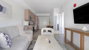A modern living room with a grey sofa, a white armchair, a coffee table, and a TV mounted on the wall. - Landing | Stylish 1BD, Gym (Hialeah)