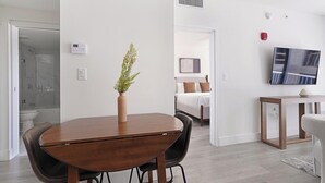 A modern hotel room with a wooden dining table, a vase with greenery, a bed with white and brown bedding, a mounted TV, and a shower area. - Landing | Stylish 1BD, Gym (Hialeah)
