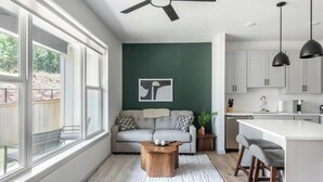 Eine moderne Küche mit weißen Schränken, ein Essbereich mit Barhocker und ein Wohnzimmer mit Sofa und Couchtisch.