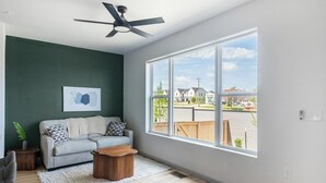 Ein Wohnzimmer mit grauem Sofa, einem Holztisch, einem Deckenventilator und einem großen Fenster mit Blick auf Häuser.