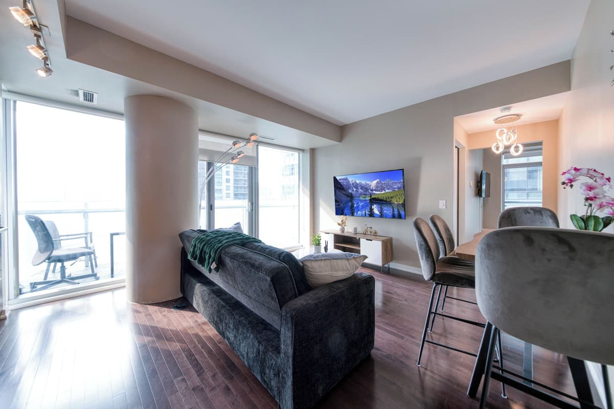 A modern living room with a sofa, a dining area with chairs, a TV mounted on the wall, and large windows allowing natural light.