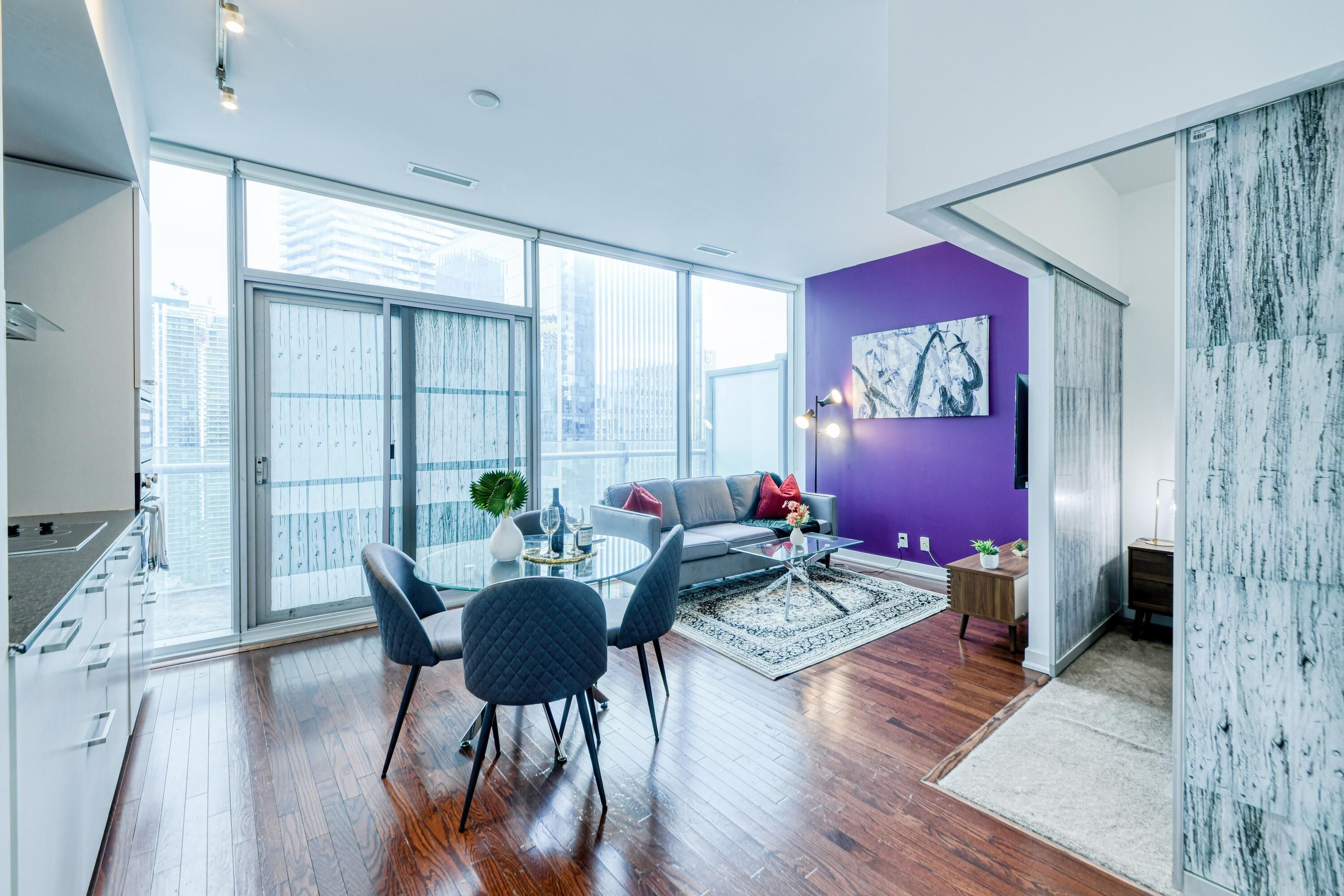 Un salon moderne avec une porte vitrée donnant sur un balcon, un mur d’accent violet et un coin repas avec une table et des chaises en verre.