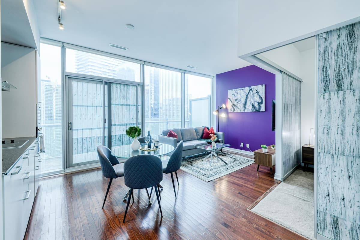 A modern living room with a glass door leading to a balcony, a purple accent wall, and a dining area with a glass table and chairs.