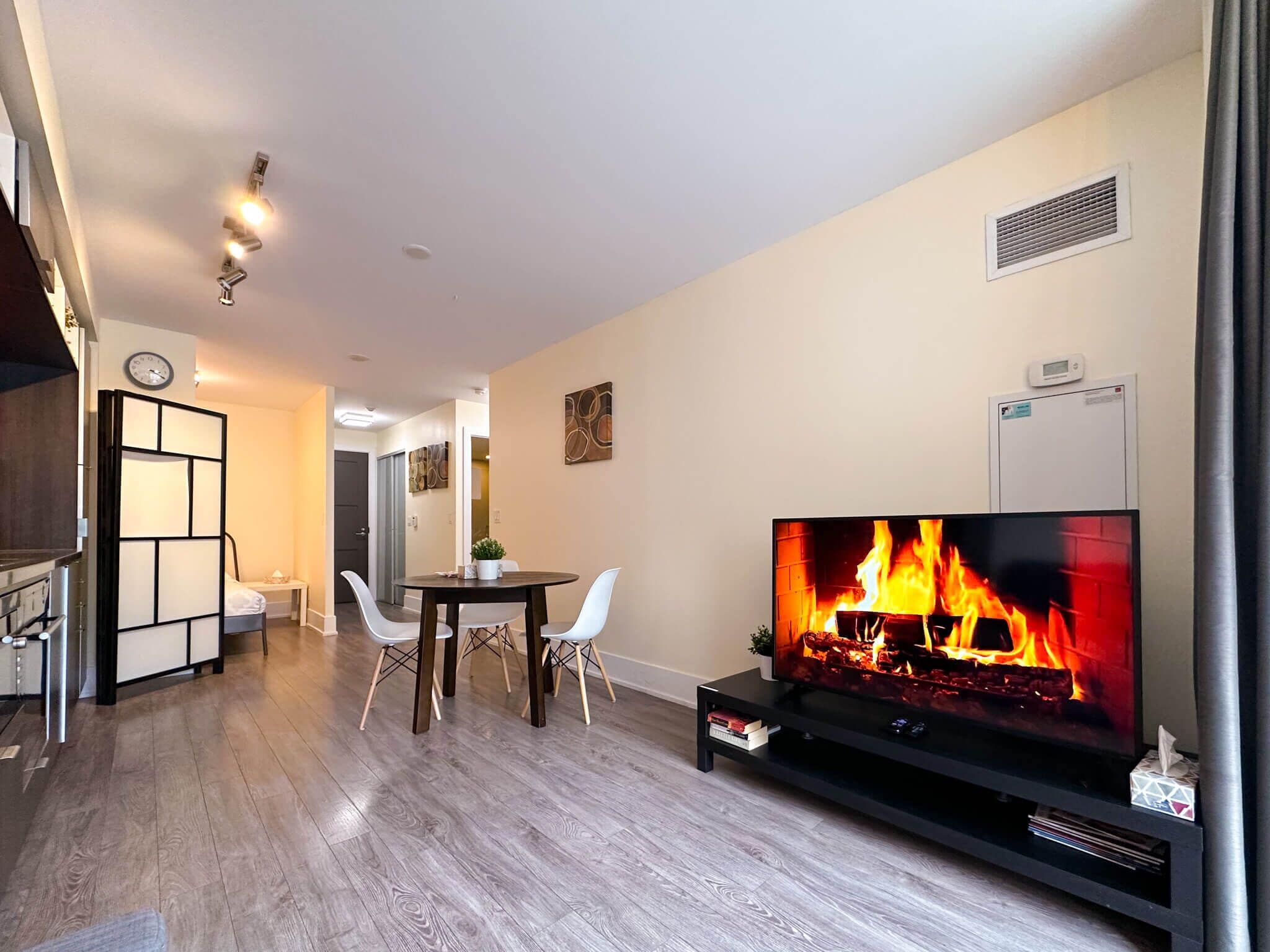 A modern living room with a flat-screen TV displaying a fireplace, a dining area with a table and chairs, and a kitchen area with cabinets and a clock.