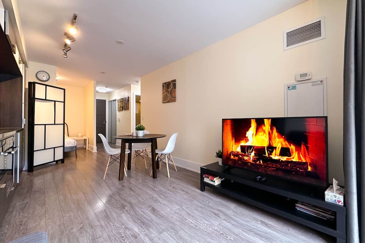 A modern living room with a flat-screen TV displaying a fireplace, a dining area with a table and chairs, and a kitchen area with cabinets and a clock.