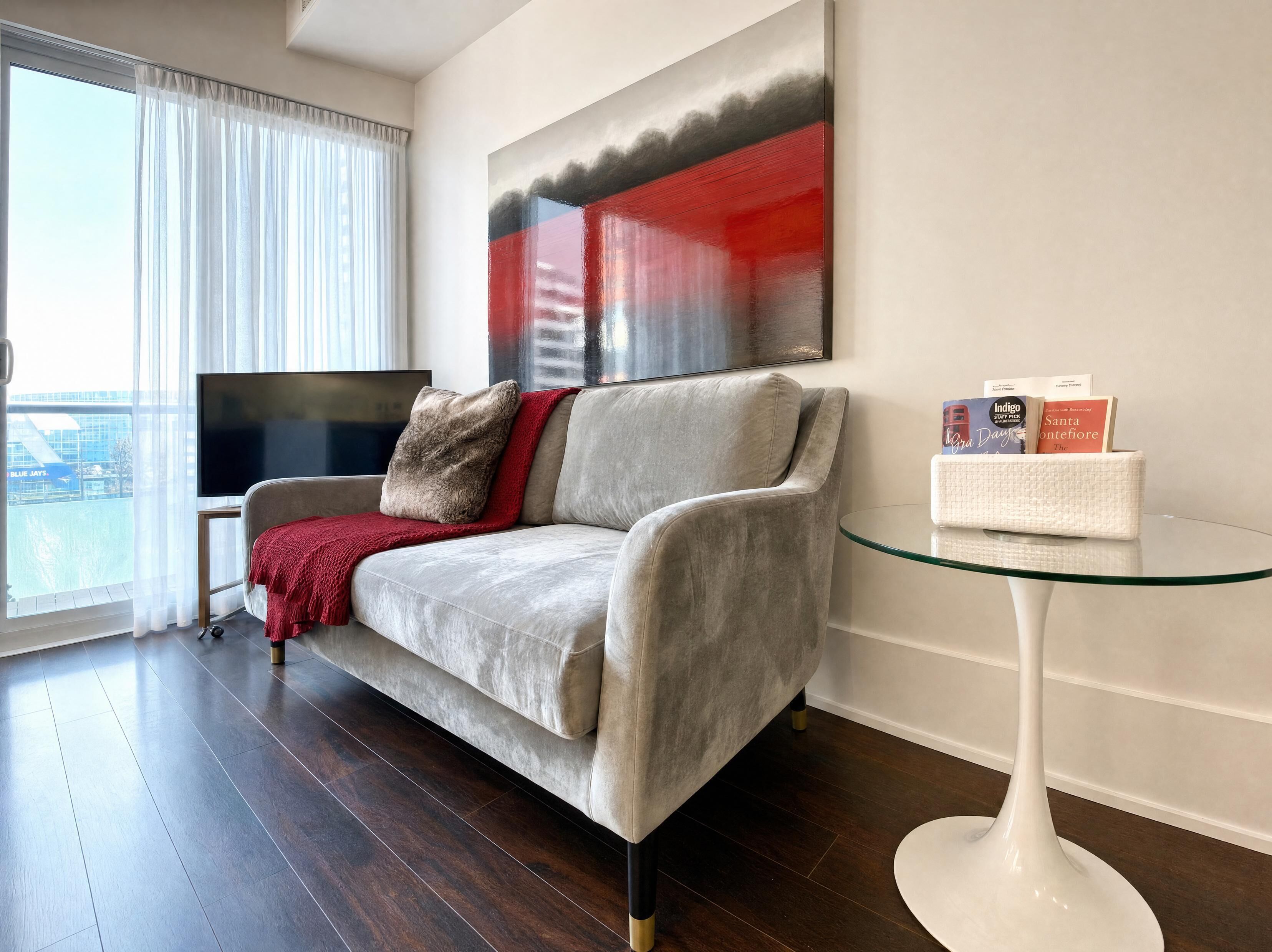 A modern living room with a grey sofa, a red throw blanket, a glass coffee table, and a flat-screen TV.