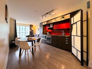 A modern kitchen with red cabinets, a dining area with a round table and chairs, and a view of a building through large windows. - Downtown Toronto Suites (Toronto)