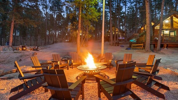 A circular fire pit surrounded by wooden Adirondack chairs in a forest setting. - Cozy 2BR Little Cabin | 15 Minutes to Mt. Rushmore (Rapid City)