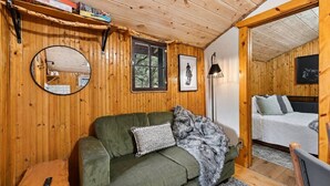 A wooden cabin interior with a green sofa, a mirror, a bed, and a small table. - Cozy 2BR Little Cabin | 15 Minutes to Mt. Rushmore (Rapid City)