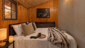 A wooden-paneled room with a bed, a nightstand, a lamp, a framed picture on the wall, and a window with a view. - Cozy 2BR Little Cabin | 15 Minutes to Mt. Rushmore (Rapid City)