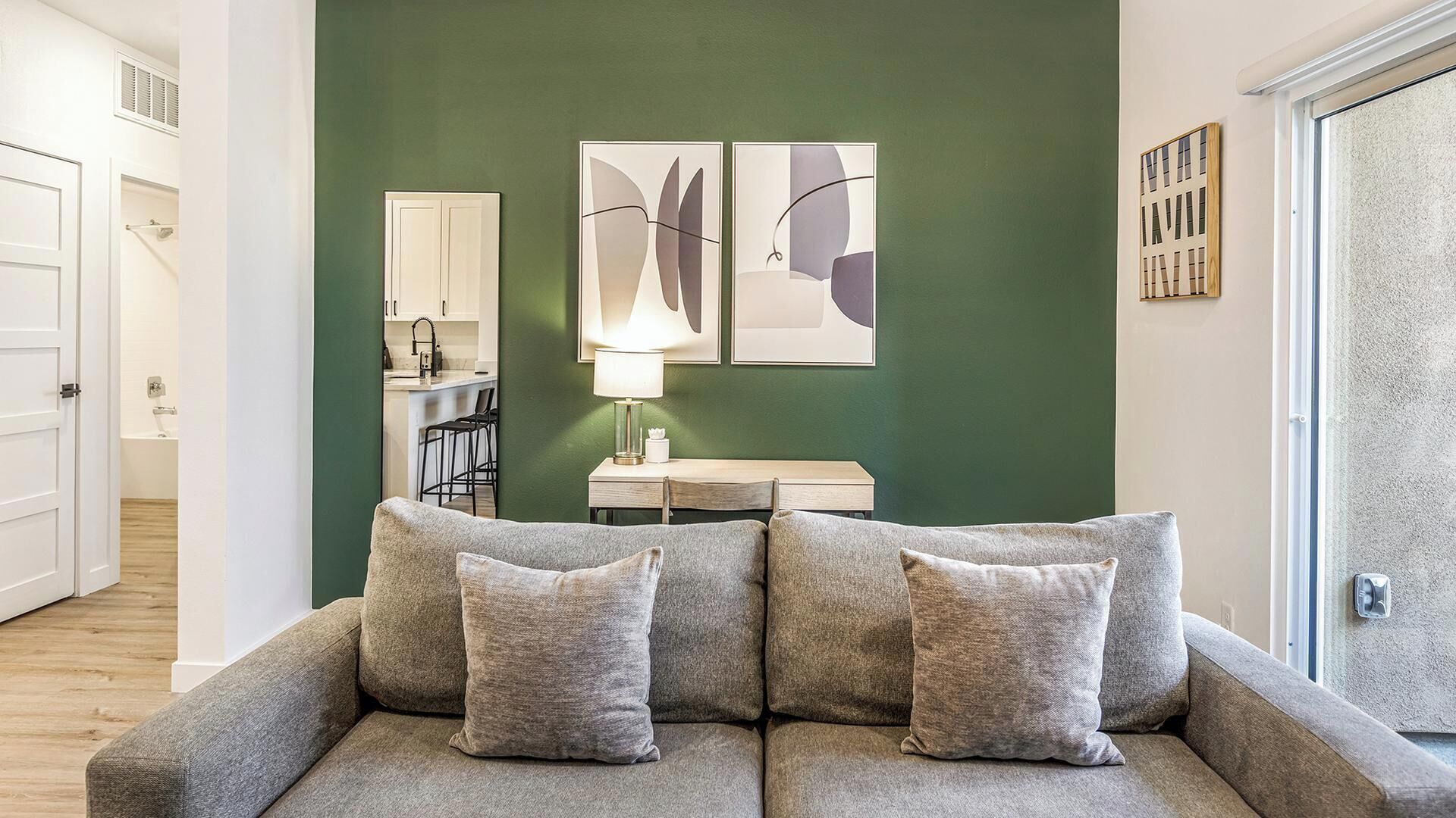 A living room with a green accent wall, a sofa with cushions, a small table with a lamp, and a kitchen area in the background.