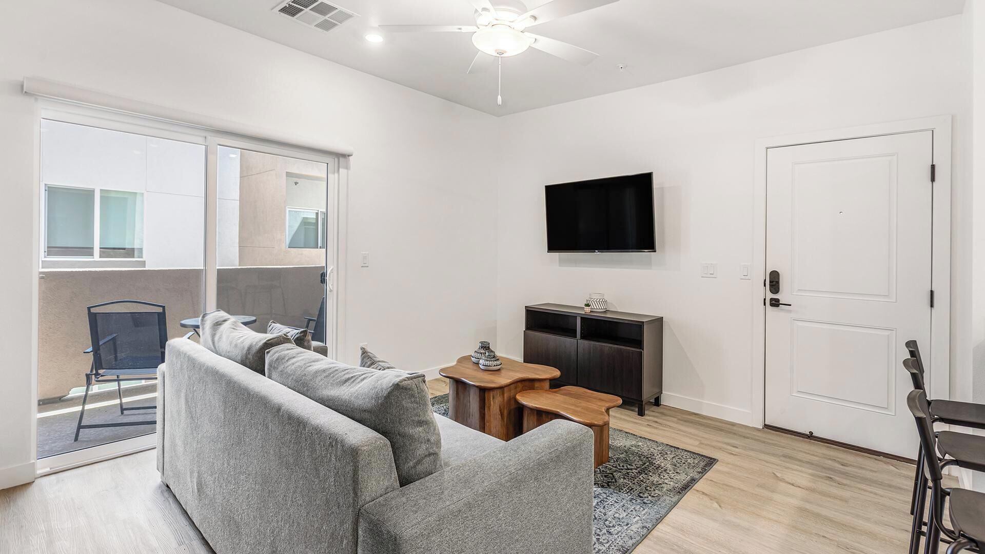 Un salon moderne avec un canapé gris, une petite table en bois, un téléviseur à écran plat fixé au mur et une porte-fenêtre coulissante donnant sur un espace extérieur.