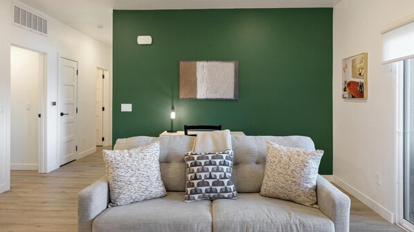 A living room with a green accent wall, a sofa with patterned cushions, a side table with a lamp, and a framed picture on the wall. - Landing | Beautiful 2BD, Clubhouse, Pool (North Las Vegas)