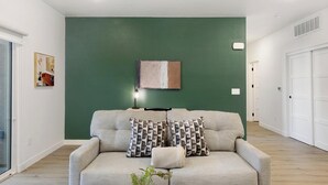Una sala de estar con una pared verde decorativa, un sofá con cojines estampados, una lámpara de pie y un cuadro enmarcado.