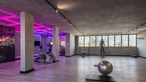 Uma academia ampla, com equipamentos de ginástica, janelas grandes e teto de concreto.