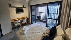 Quarto de hotel com uma cama, uma escrivaninha com cadeira, televisão e varanda com vista para edifícios.
