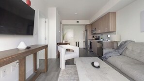 A modern living room with a sofa, a coffee table, a TV mounted on the wall, and a kitchen area with cabinets and appliances. - Landing | Amazing 1BD, Gym (Hialeah)
