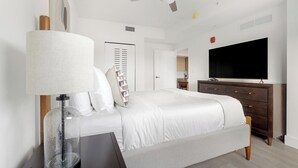 A bedroom with a bed, a bedside table, a lamp, a dresser, a TV, and a door. - Landing | Chic 1BD, Gym (Hialeah)