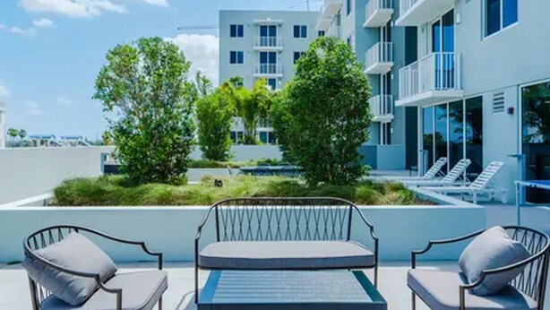 A rooftop terrace with a table and chairs, a swimming pool, and a modern building in the background. - Landing | Chic 1BD, Gym (Hialeah)