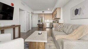 A modern living room with a sofa, coffee table, TV, and kitchen in the background. - Landing | Chic 1BD, Gym (Hialeah)